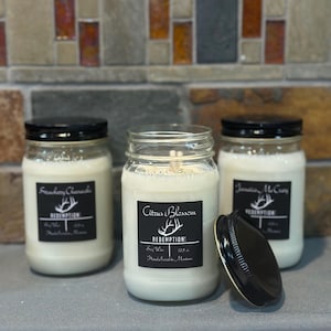 May include: Three glass jar candles with black lids. The candles have white wax and black labels with white text. The labels read "Strawberry Cherish", "Citrus Blossom", and "Jamaica McCray". The candle jar lids are black.