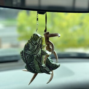 May include: A car air freshener featuring a detailed bass fish design in shades of green and black, accented with natural-colored antler elements. The air freshener hangs from a rearview mirror, adding a touch of nature to the vehicle's interior.