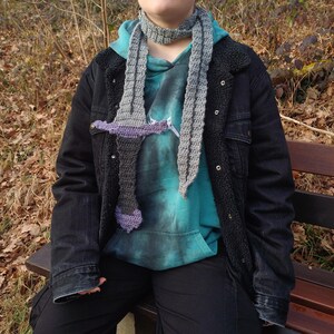 Könnte beinhalten: Handgehäkelter Schal in Grau- und Lilatönen, der einem Schwert nachempfunden ist. Der Schal wird über einem türkisfarbenen Kapuzenpullover und einer schwarzen Jeansjacke getragen. Das Design des Schals umfasst einen Parier und eine Klinge.
