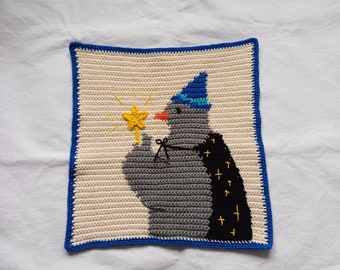 May include: Crocheted square featuring a gray bird wearing a blue wizard hat and black cape with yellow stars. The bird holds a yellow star wand. The square has a cream background with a blue border.