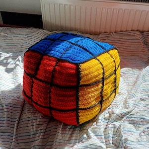 May include: A handmade, cube-shaped pouf in a Rubik's Cube design. The crocheted pouf features red, yellow, and blue squares separated by black lines. The cube-shaped pouf is a decorative and functional item for a living room or bedroom.