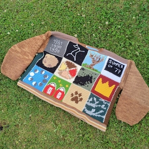 May include: A handmade, patchwork crochet sweater with brown sleeves and a multi-colored body. The body features various square designs, including text, a paw print, a crown, and a constellation. The text "SUMMER OF '77" is visible on one square.