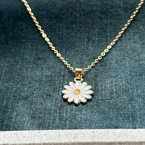 Könnte beinhalten: Eine goldene Kettenkette mit einem weißen Gänseblümchen-Anhänger. Die Blume hat eine gelbe Mitte.
