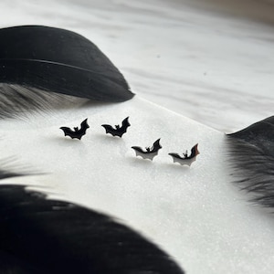 Könnte beinhalten: Vier kleine, schwarze Fledermaus-Ohrringe sind auf einer weißen Oberfläche angeordnet, mit zwei großen schwarzen Federn auf beiden Seiten. Die Ohrringe haben ein schlichtes, minimalistisches Design, perfekt für Halloween oder den Alltag. Das Bild hat eine klare, moderne Ästhetik.