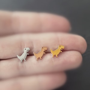 Könnte beinhalten: Drei Dinosaurier-Ohrringe in Silber, Gold und Roségold. Die Dinosaurier sind im Pixel-Stil gehalten, ähnlich dem klassischen Videospielcharakter.