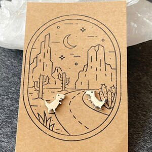 Puede incluir: Un par de pendientes de plata con un dise&ntilde;o de dinosaurio pixelado. Los pendientes se muestran en una tarjeta marr&oacute;n con una ilustraci&oacute;n en blanco y negro de un paisaje des&eacute;rtico con cactus y una luna creciente.