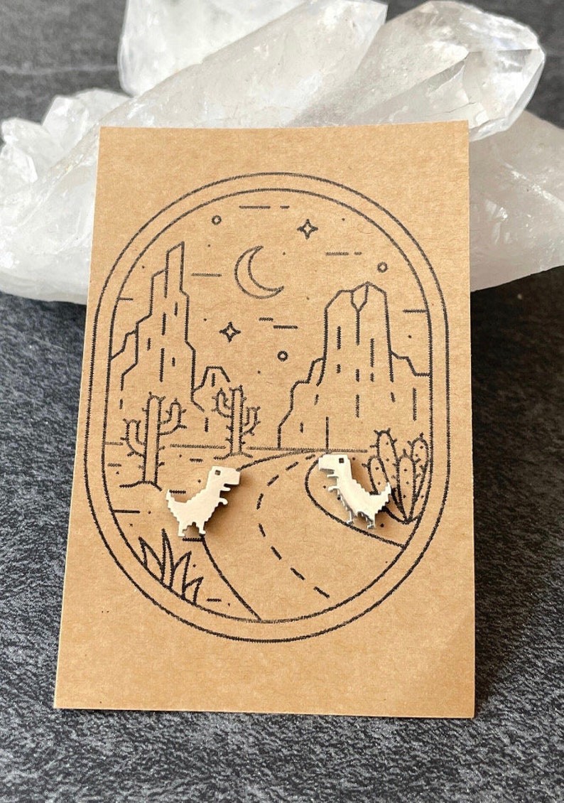 Puede incluir: Un par de pendientes de plata con un dise&ntilde;o de dinosaurio pixelado. Los pendientes se muestran en una tarjeta marr&oacute;n con una ilustraci&oacute;n en blanco y negro de un paisaje des&eacute;rtico con cactus y una luna creciente.