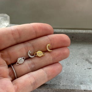 Könnte beinhalten: Eine Sammlung von Ohrsteckern zum Thema Himmel. Die Ohrringe umfassen einen silbernen Planeten mit Kristallmuster, einen silbernen Halbmond, einen goldenen Planeten und einen goldenen Halbmond. Die Ohrringe sind klein und zart.
