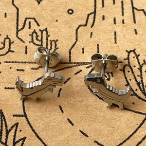 Puede incluir: Un par de pendientes de plata con forma de dinosaurio. Los pendientes son peque&ntilde;os y detallados, con un acabado texturizado.