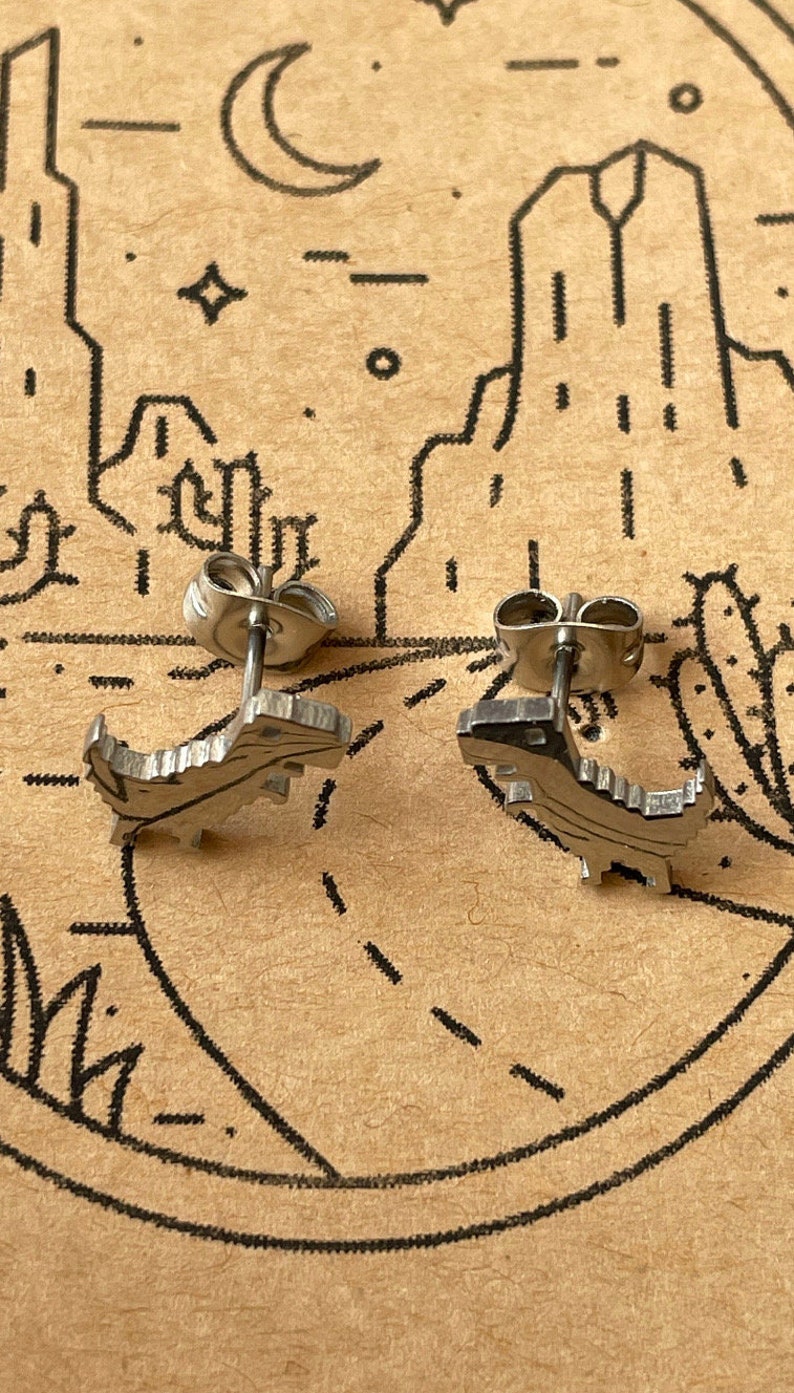 Puede incluir: Un par de pendientes de plata con forma de dinosaurio. Los pendientes son peque&ntilde;os y detallados, con un acabado texturizado.