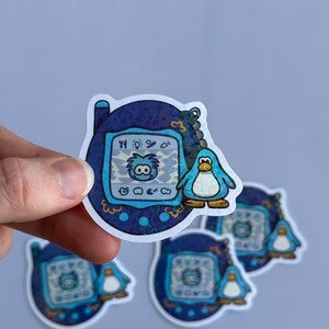 Op de afbeelding: Een blauw en wit digitaal huisdier speelgoed met een pingu&iuml;n karakter op het scherm. Het speelgoed is omgeven door een blauwe en witte gepatroneerde rand. Het speelgoed is een sticker.
