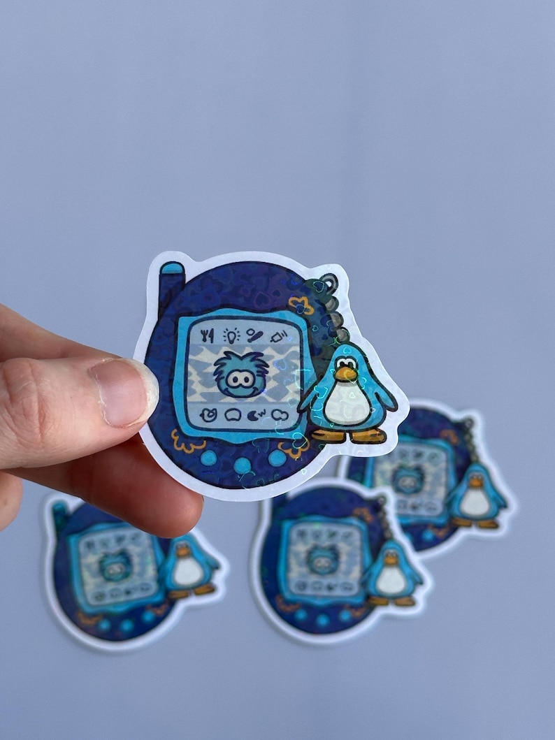 Op de afbeelding: Een blauw en wit digitaal huisdier speelgoed met een pingu&iuml;n karakter op het scherm. Het speelgoed is omgeven door een blauwe en witte gepatroneerde rand. Het speelgoed is een sticker.