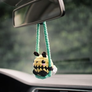 Puede incluir: Una abeja de crochet colgando del espejo retrovisor de un coche. La abeja es amarilla y negra con una cara blanca y mejillas rosadas.