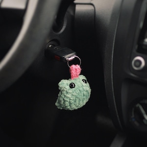 以下が含まれることがあります： 車のイグニッションに付いている車のキーに、黒い目とピンクのハートがついた緑色の編みぐるみがぶら下がっています。