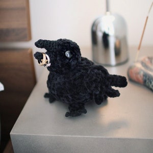 May include: A black crocheted crow with large eyes holding two gold rings in its beak. The crow is sitting on a gray surface.