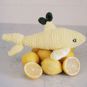 May include: A crocheted yellow lemon-shaped plush toy with green leaves on top, surrounded by real lemons.