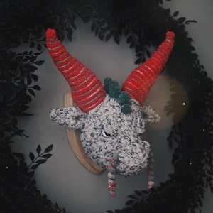 May include: A gray and white crocheted dragon head with red and white striped horns and green accents. The dragon head is mounted on a wooden plaque and has candy cane-like decorations hanging from its mouth.