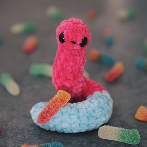 May include: A pink and blue crocheted worm toy with black eyes. The worm is surrounded by colorful gummy worms.