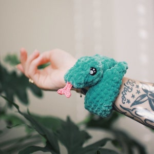 May include: A teal green crocheted snake bracelet with a pink tongue and black eyes. The bracelet is worn on a person's arm.