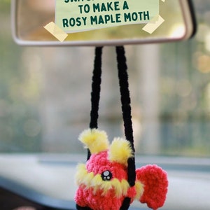 May include: A crocheted rosy maple moth hanging from a car's rearview mirror. The moth is pink and yellow with black yarn for the hanging strings. A yellow sticky note on the mirror reads "Switch colours to make a rosy maple moth."