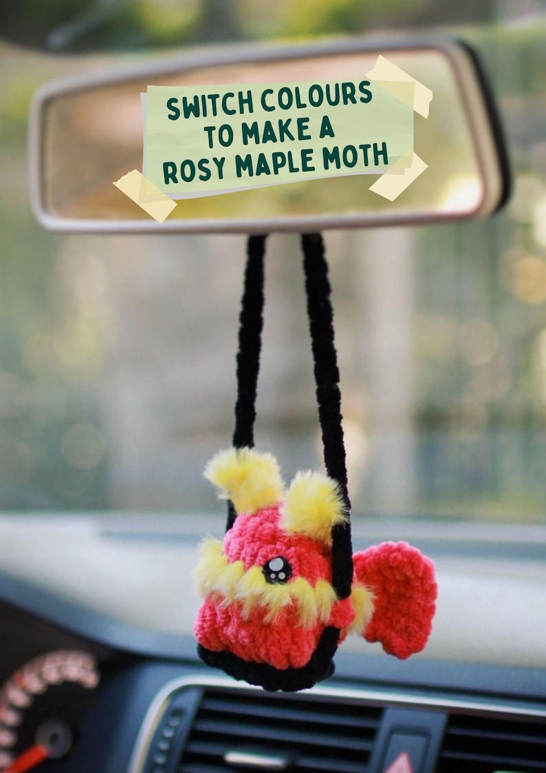 May include: A crocheted rosy maple moth hanging from a car's rearview mirror. The moth is pink and yellow with black yarn for the hanging strings. A yellow sticky note on the mirror reads "Switch colours to make a rosy maple moth."