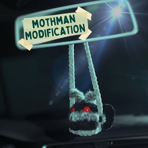 May include: A grey and black fuzzy creature with red eyes hangs from the rear-view mirror of a car. The mirror has a sign that reads "Mothman Modification."