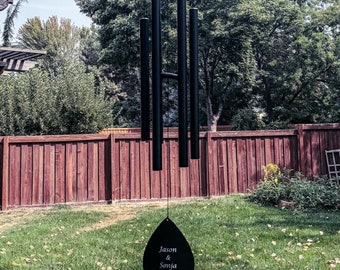 May include: Black metal wind chime with a teardrop-shaped charm that reads "Jason & Sonja" with a heart symbol. The chime is hanging from a wooden pergola.