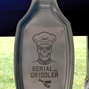 May include: A stainless steel spatula holder with a skull and chef hat design. The holder is engraved with the text "Jason Serial Griddler" and a small spatula icon.