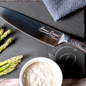 Puede incluir: Un cuchillo de chef de acero inoxidable con mango de madera, grabado con el texto "Jason Deines", descansa sobre una tabla de cortar de pizarra negra. Tambi&eacute;n hay esp&aacute;rragos frescos y un cuenco peque&ntilde;o con polvo blanco.