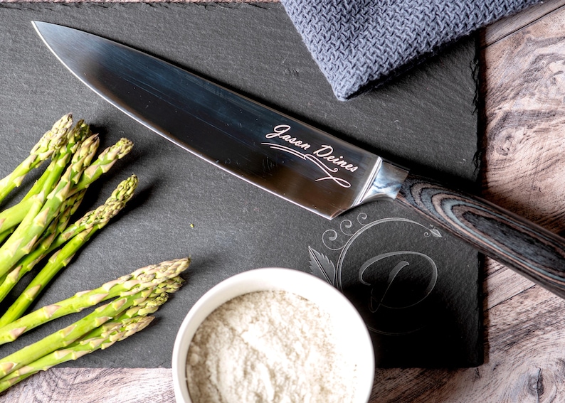 Puede incluir: Un cuchillo de chef de acero inoxidable con mango de madera, grabado con el texto "Jason Deines", descansa sobre una tabla de cortar de pizarra negra. Tambi&eacute;n hay esp&aacute;rragos frescos y un cuenco peque&ntilde;o con polvo blanco.
