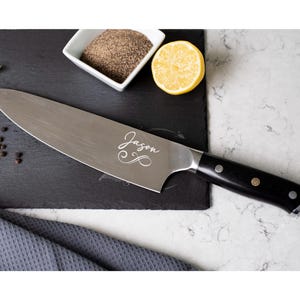 May include: A stainless steel chef's knife with a black handle secured by three metal rivets. The blade features the engraved name Jason in a cursive script with a decorative swirl flourish underneath the text, positioned on a dark slate surface.