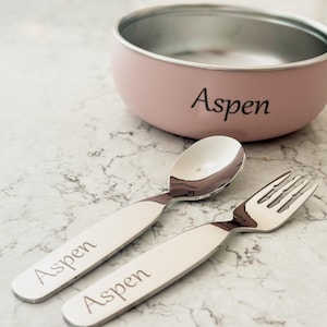 May include: A pink and silver pet bowl with matching spoon and fork. The bowl and utensils have the name "Aspen" engraved on them. The set is designed for a pet.