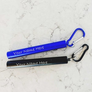 May include: Two personalized metal straws with attached keychains. One straw is blue and the other is black. Both straws have the text "Your Name Here" engraved on them.