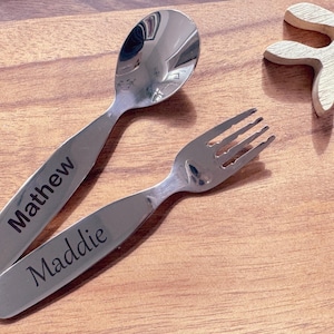 May include: A set of two personalized stainless steel utensils, a spoon and a fork. The spoon is engraved with the name "Matthew" and the fork is engraved with the name "Maddie".