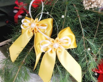 Könnte beinhalten: Zwei gelbe Satin-Schmuckbänder mit weißen Blumenmitten und Perlenakzenten. Die Schleifen haben lange, fließende Bänder und sind an weißen Schlaufen aufgehängt. Die Ornamente sind vor Tannenzweigen und anderen festlichen Dekorationen platziert.