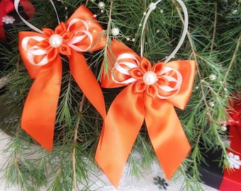 Könnte beinhalten: Zwei leuchtend orangefarbene Satin-Schleifen mit weißen Bändern und Perlen in der Mitte. Sie sind vor einem Hintergrund aus grünen Tannennadeln und weißen Perlen platziert, passend für Feierlichkeiten.