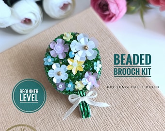 Könnte beinhalten: Ein Perlenbroschen-Set mit floralem Design. Die Brosche zeigt weiße, gelbe und lila Blumen mit grünen Blättern. Der Text "Beaded Brooch Kit" und "Beginner Level" sind sichtbar.