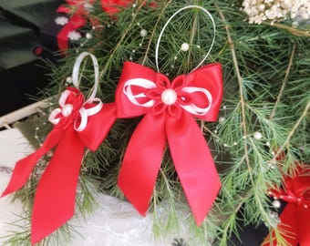 Könnte beinhalten: Zwei rote Satin-Schleifen mit weißen Akzenten und Perlenverzierungen. Die Schleifen hängen an weißen Schlaufen vor einem Hintergrund aus Tannenzweigen und weißen floralen Akzenten. Festliche Weihnachtsdekoration.