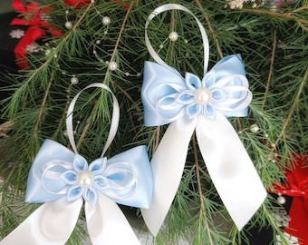 Könnte beinhalten: Zwei hellblaue und weiße Satin-Schleifen-Ornamente mit Perlenakzenten. Die Schleifen haben eine blütenförmige Mitte und lange weiße Bänder. Sie werden auf einem Bett aus grünen Tannennadeln mit roten und schwarzen Geschenkboxen im Hintergrund präsentiert.