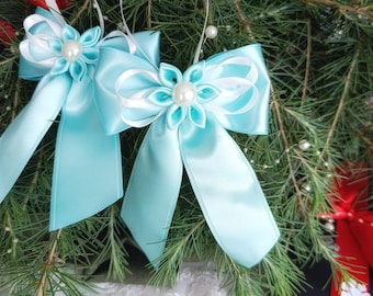 Könnte beinhalten: Zwei hellblaue Satin-Schleifen mit weißen Akzenten und Perlen in der Mitte. Die Schleifen sind an weißen Bändern befestigt und vor dem Hintergrund von Tannenzweigen und roten Geschenkboxen platziert. Dekorative Schleifen für Feiertagsdekoration.