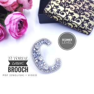 May include: A silver beaded brooch in the shape of the letter C. The brooch is on a white background with a pink flower and a black box with a gold design. The text "TUTORIAL Letter C BROOCH PDF (ENGLISH) + VIDEO" is below the brooch. The text "BEGINNER LEVEL" is in a circle above the brooch.