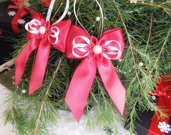 Könnte beinhalten: Zwei leuchtend rosa Satinband-Weihnachtsschmuckstücke mit weißen Akzenten und Perlen in der Mitte hängen an weißen Schlaufen. Vor dem Hintergrund von Tannenzweigen und kleinen weißen Perlen, mit verpackten Geschenken im Hintergrund.