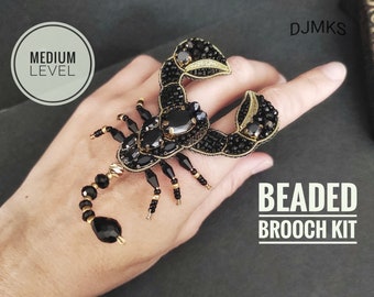 Könnte beinhalten: Schwarzes, perlenbesetztes Skorpion-Broschen-Set. Der Skorpion ist mit schwarzen Perlen und Edelsteinen mit goldenen Akzenten verziert. Der Text "BEADED BROOCH KIT" wird in Weiß angezeigt. Der Text "MEDIUM LEVEL" befindet sich in einem weißen Kreis.