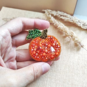 Puede incluir: Un broche de calabaza con cuentas, con un tallo verde y cuentas naranjas y transparentes.