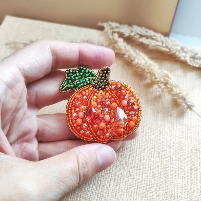 Puede incluir: Un broche de calabaza con cuentas, con un tallo verde y cuentas naranjas y transparentes.