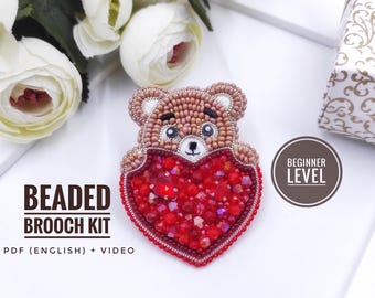 Könnte beinhalten: Ein Perlenbroschen-Set mit einem braunen Teddybären, der ein rotes Herz hält. Der Bär besteht aus kleinen Perlen, und das Herz ist mit roten Kristallen gefüllt. Der Text "BEADED BROOCH KIT" wird angezeigt. Das Set ist mit "BEGINNER LEVEL" gekennzeichnet.