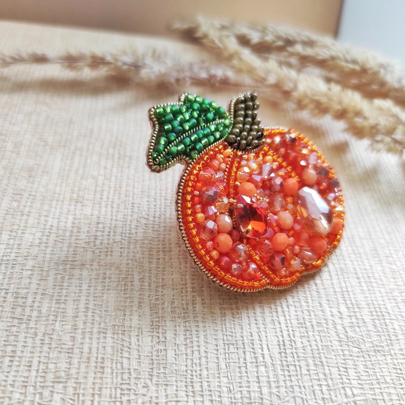 Puede incluir: Un broche de calabaza con cuentas, con un tallo verde y cuentas naranjas y doradas.