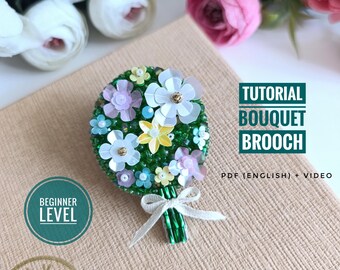 Könnte beinhalten: Handgefertigte Brosche in Form eines Blumenstraußes mit weißen, gelben und lila Blüten mit goldenen Zentren, umgeben von grünen Perlen. Die Brosche ist mit einem weißen Band gebunden. Der Text auf dem Bild lautet "Tutorial Bouquet Brooch" und "Beginner Level".