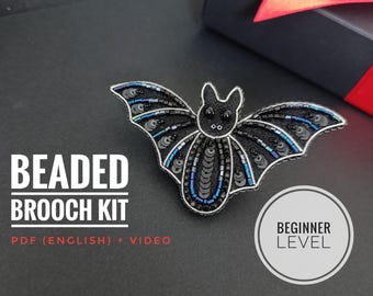 Könnte beinhalten: Ein besticktes Fledermaus-Broschen-Set mit schwarzen Pailletten und blauen Akzenten. Die Brosche hat eine silberne Kontur und liegt auf einer schwarzen Oberfläche. Der Text auf dem Bild lautet "Beaded Brooch Kit" und "Beginner Level".