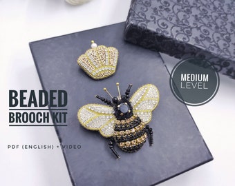 Könnte beinhalten: Ein Perlenbroschen-Set mit einem Bienen- und Kronen-Design. Die Bienenbrosche hat schwarze, goldene und weiße Perlendetails mit einem schwarzen Edelstein. Die Krone ist gold- und weißperlenbesetzt. Der Text "BEADED BROOCH KIT" wird angezeigt.
